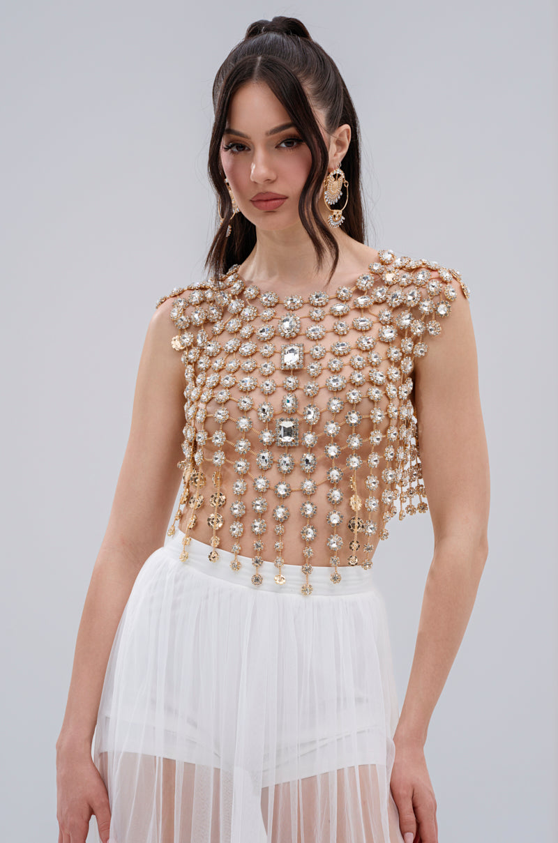 A woman with long dark hair models the BEJEWELED GOLD SLEEVELESS TOP, featuring gemstone embellishments, over a white skirt. She wears large ornate earrings and poses against a light gray background.
