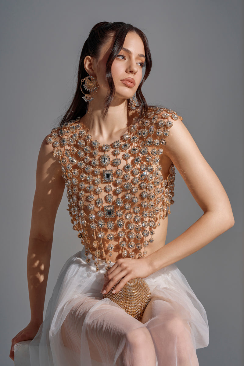 A woman with long brown hair poses in the BEJEWELED GOLD SLEEVELESS TOP, its sparkling gemstone embellishments catching the light. She pairs it with a white tulle skirt and large earrings, gazing to the side against a neutral background.