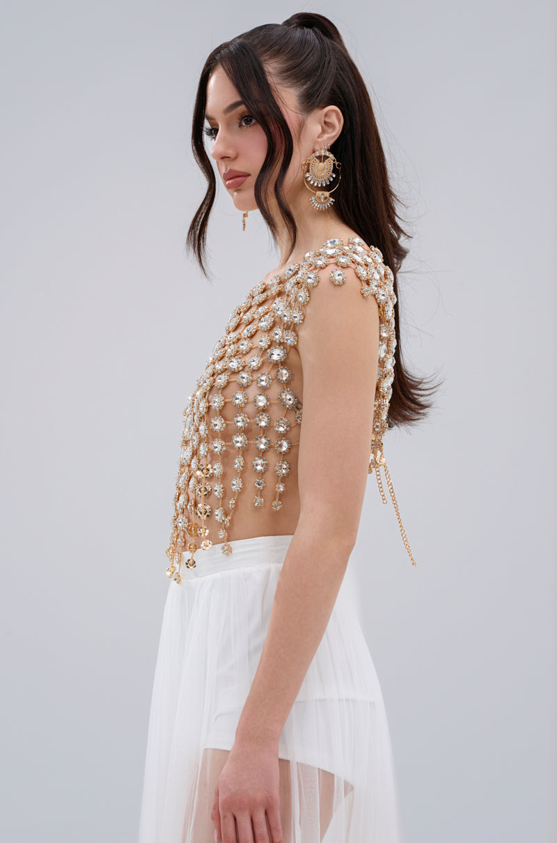A woman with long dark hair in a high ponytail wears the BEJEWELED GOLD SLEEVELESS TOP with pearl and gemstone accents, paired with a white sheer asymmetrical skirt and large ornate earrings, standing against a plain background.