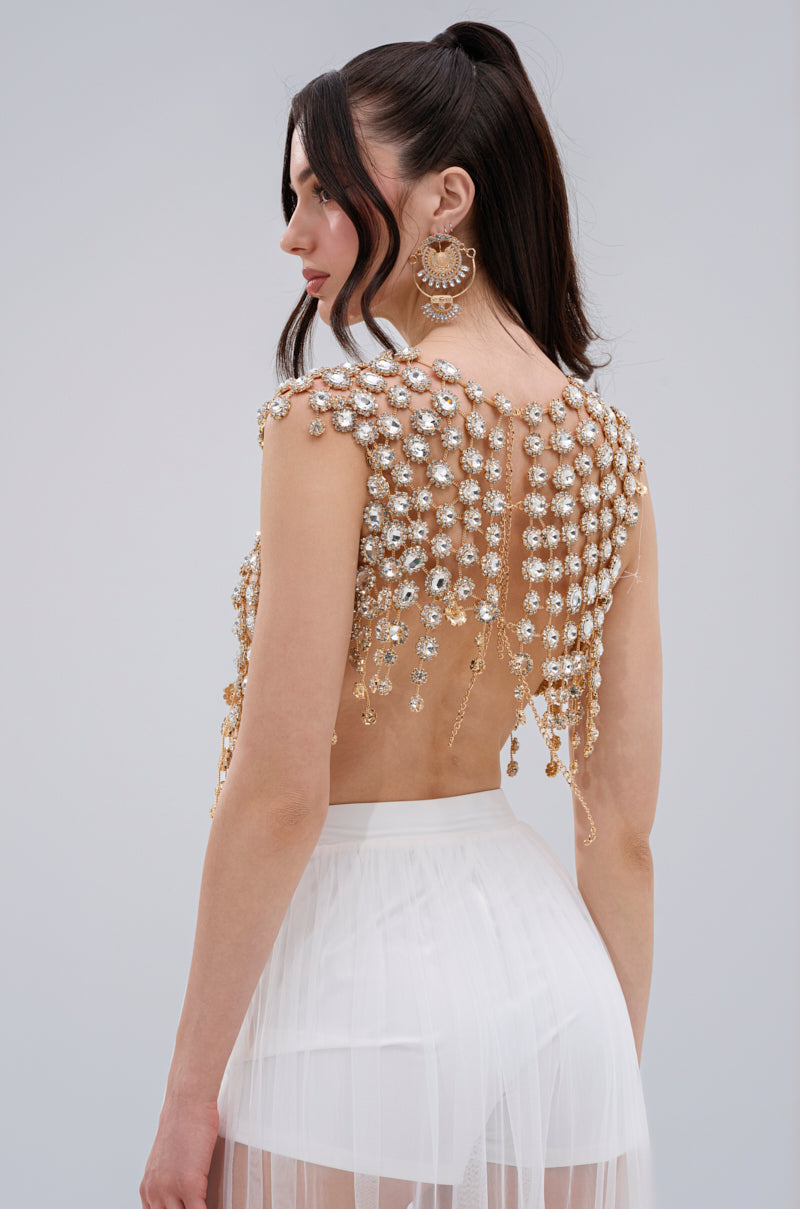 A woman with long dark hair in a high ponytail models the BEJEWELED GOLD SLEEVELESS TOP with gemstone embellishments, paired with a sheer white skirt and large statement earrings, glancing over her shoulder.