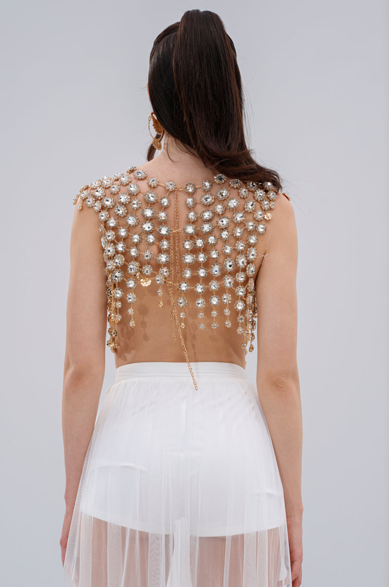 With their back to the camera, a person wears the BEJEWELED GOLD SLEEVELESS TOP with gemstones and a white sheer skirt. Their long, dark hair is partially tied back against a plain, light-colored background.