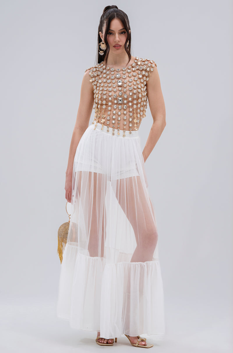 A woman wears the BEJEWELED GOLD SLEEVELESS TOP, paired with a sheer white floor-length skirt, gold heels, a small gold fringed bag, and straight hair.