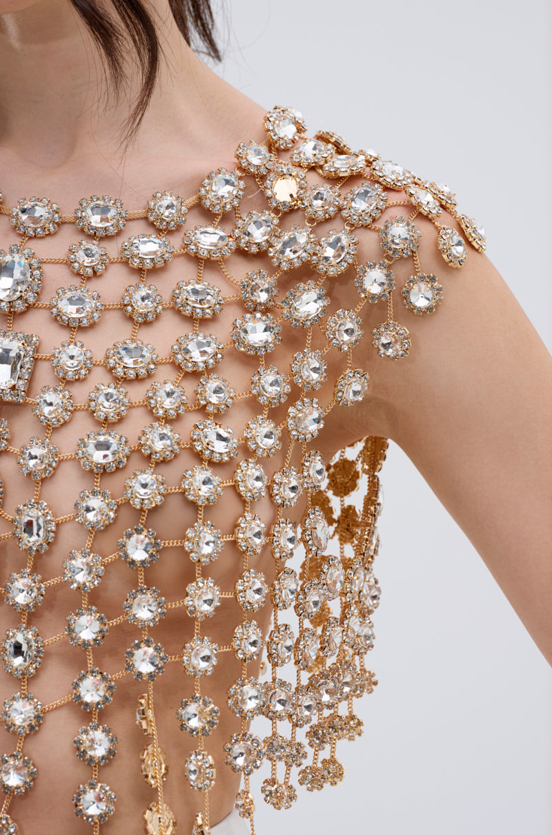 A close-up of a person wearing the BEJEWELED GOLD SLEEVELESS TOP, featuring metallic chain links and large sparkling gemstones draped over the shoulder and chest, against a plain light background.