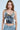 A person with long brown hair wears a BLING BLING TOP, adorned in shiny sequin discs, and light blue jeans against a matching background. Their expression is neutral, with hands tucked into jean pockets—perfect for a party.