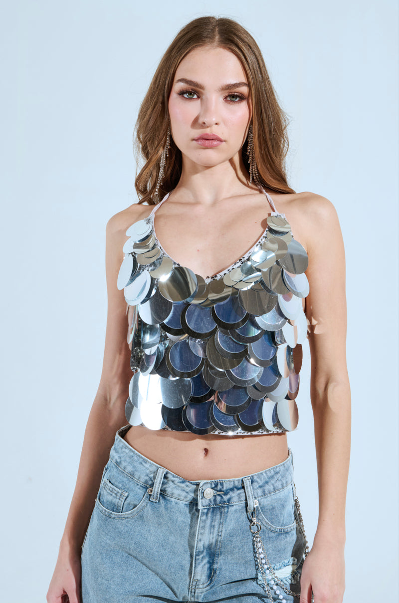 A woman stands against a simple backdrop, wearing the party-ready BLING BLING TOP paired with light blue jeans. Her hair falls freely, decorated with long earrings. She exudes confidence as the sequin discs of her top shimmer brilliantly.