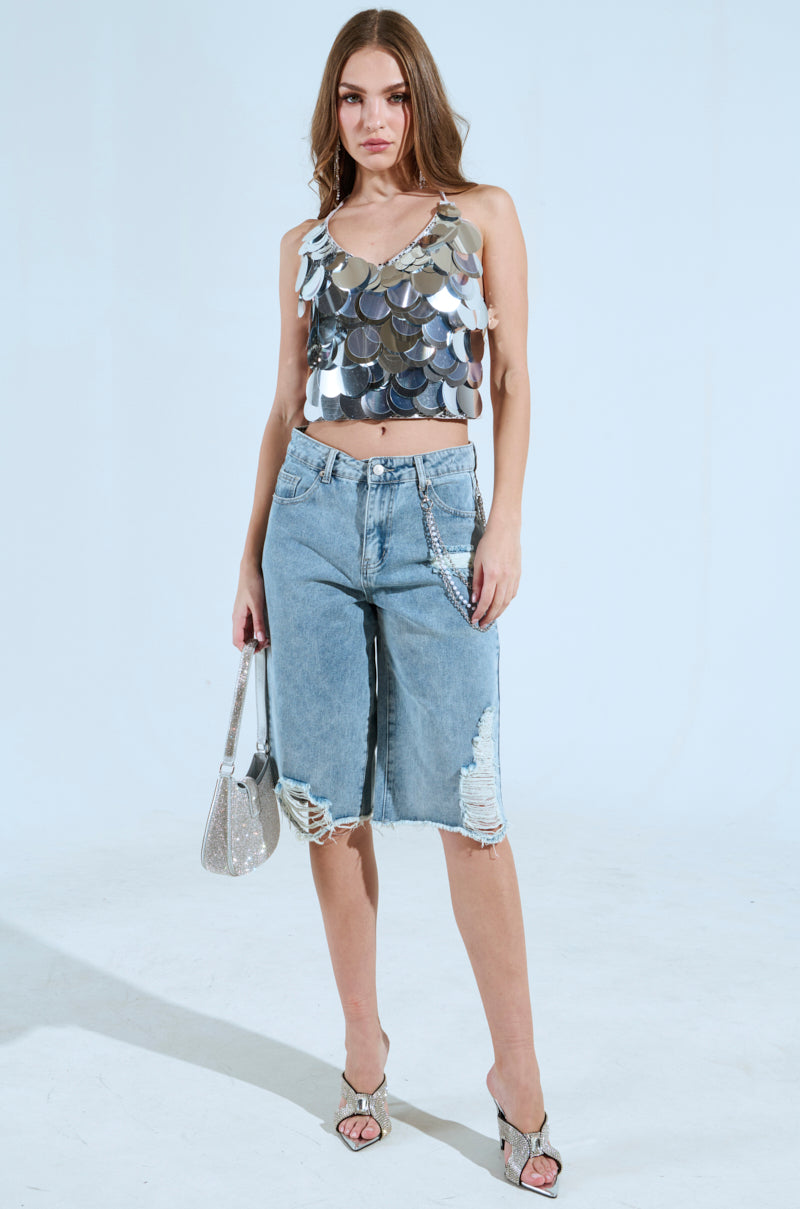 A woman poses in a BLING BLING TOP with large sequin discs, paired with distressed denim shorts and heels. She holds a silver handbag, her long straight hair flowing against a plain light background.