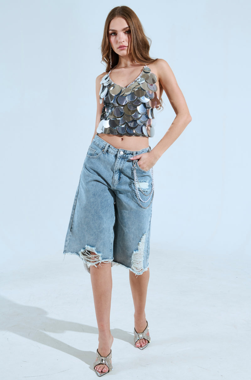 A woman confidently poses against a plain background, showcasing the BLING BLING TOP with sequin discs and faded denim shorts featuring distressed details. Her party-perfect look is completed with long wavy hair and heeled sandals.
