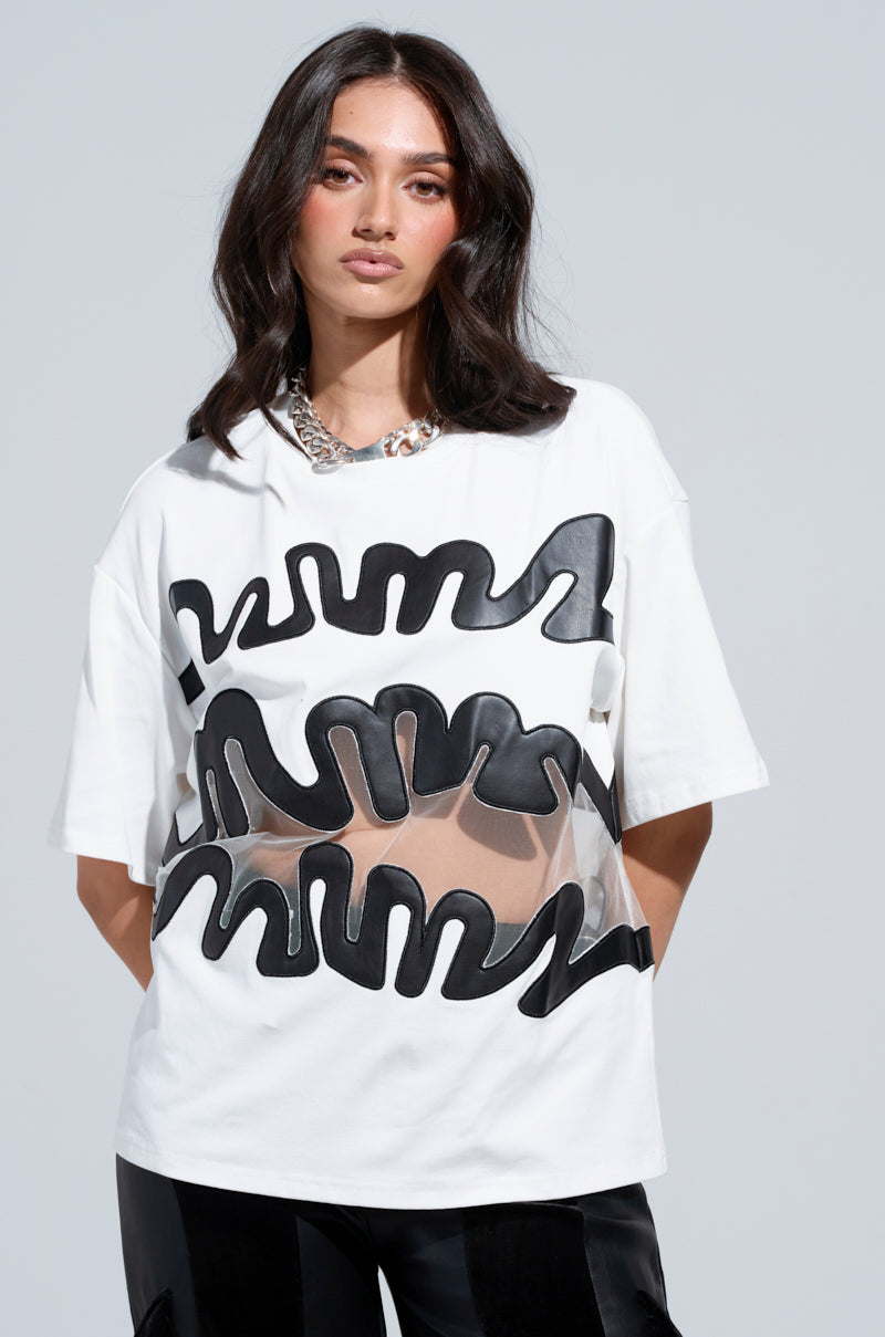 A person with long dark hair is wearing the MIND THE GAP MESH TRIM SHIRT, featuring bold black wavy patterns and sheer elements. They have a neutral expression and are accessorized with a chunky silver chain necklace, set against a plain gray background.