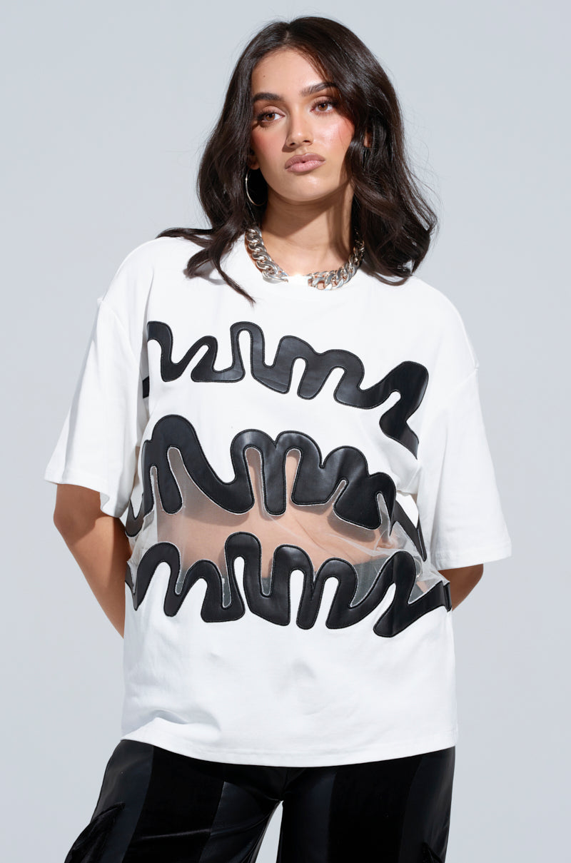 A woman wears the MIND THE GAP MESH TRIM SHIRT, a white t-shirt adorned with bold black abstract designs and featuring a sheer mesh cutout panel at the center. Her long dark hair flows elegantly, enhanced by large hoop earrings and a chunky silver chain necklace. The plain, neutral background accentuates her chic style.