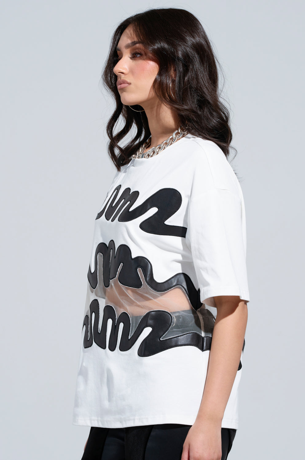 A woman with long, dark hair models the MIND THE GAP MESH TRIM SHIRT with abstract faux leather accents and sheer mesh cutout panels. She complements her look with a silver chain necklace and is photographed from the side against a simple background.