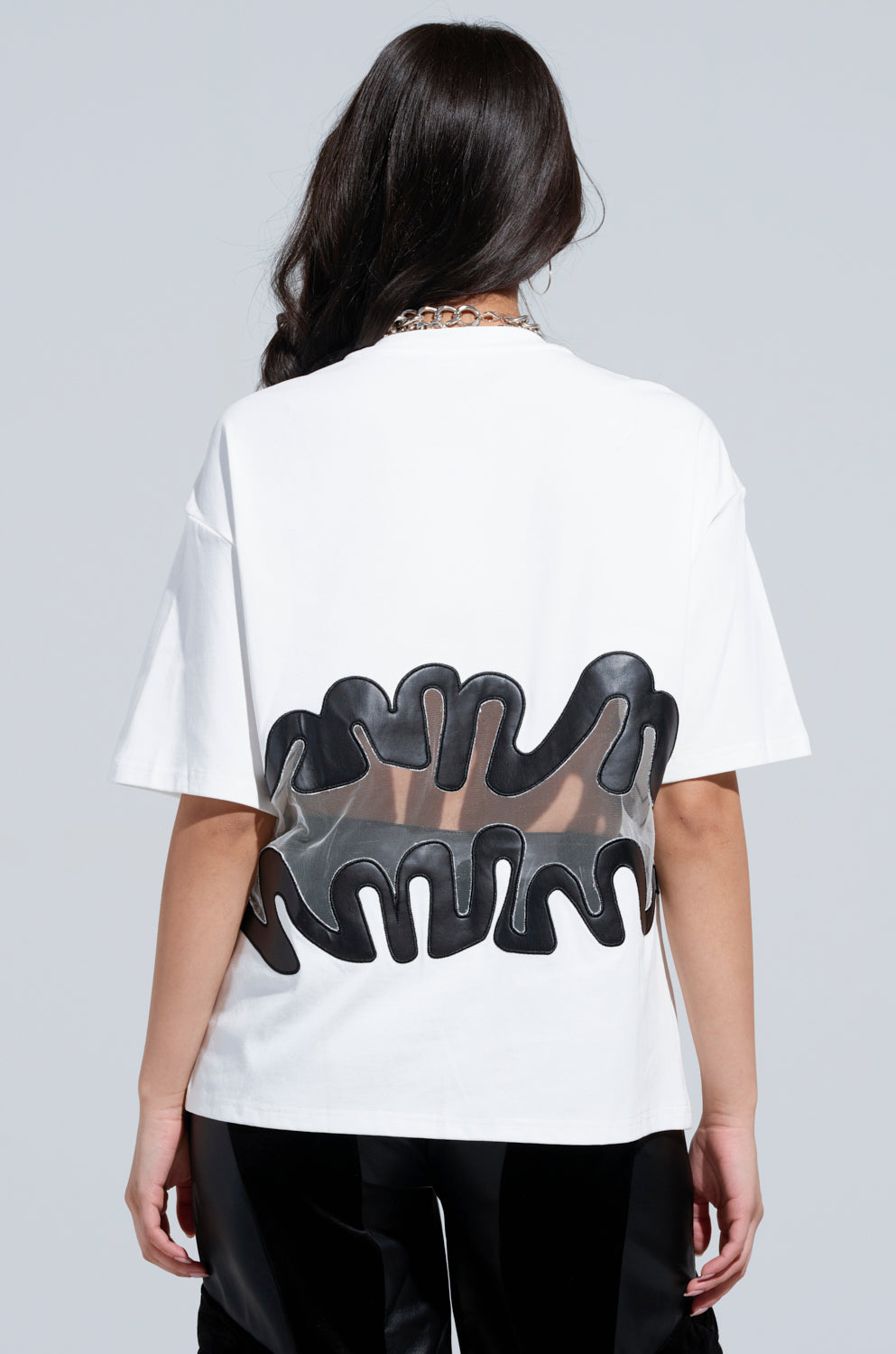 A person wearing the MIND THE GAP MESH TRIM SHIRT, which features a distinctive black wavy pattern and a sheer mesh cutout panel on the back. The individual has long, dark hair and accessorizes with a silver chain necklace, set against a plain gray background.