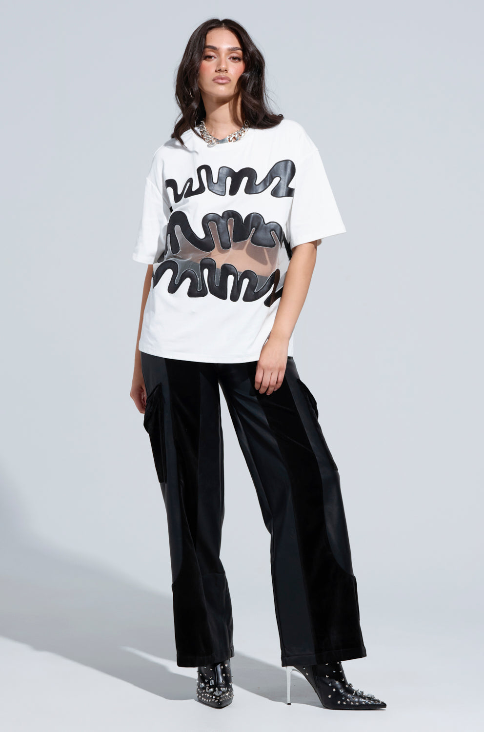 A person poses confidently in the MIND THE GAP MESH TRIM SHIRT, characterized by bold black abstract designs, paired with black shiny pants. They complete the look with studded black ankle boots and have long hair, set against a plain light gray backdrop.