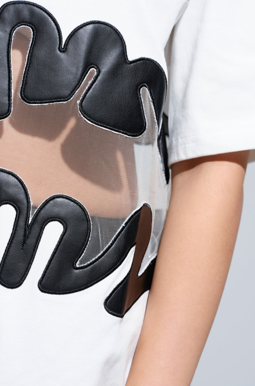 Close-up of a person's torso showcasing the MIND THE GAP MESH TRIM SHIRT in white, featuring abstract faux leather trim and sheer mesh cutout panels. The design forms a wavy, artistic pattern on the fabric, with a plain background that highlights the shirt's intricate details.