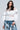 A person with long, dark hair stands confidently wearing the CUT HIM OFF SWEATSHIRT IN IVORY, featuring textured sheer panels that give a stylish look. Complementing the outfit are loose-fit jeans decorated with black cross designs and an elegant chain necklace with an ornate pendant.