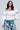 A person with long dark hair is wearing the CUT HIM OFF SWEATSHIRT IN IVORY, accented with sheer mesh panels and a silver cross necklace. They are paired with blue jeans adorned with black and white cross patterns, set against a simple, light-colored background.