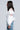 A person with wavy dark hair is wearing the CUT HIM OFF SWEATSHIRT IN IVORY, which features transparent stripe panels, paired with blue jeans embellished with white floral designs. The cropped silhouette stands against a light gray background, facing sideways.