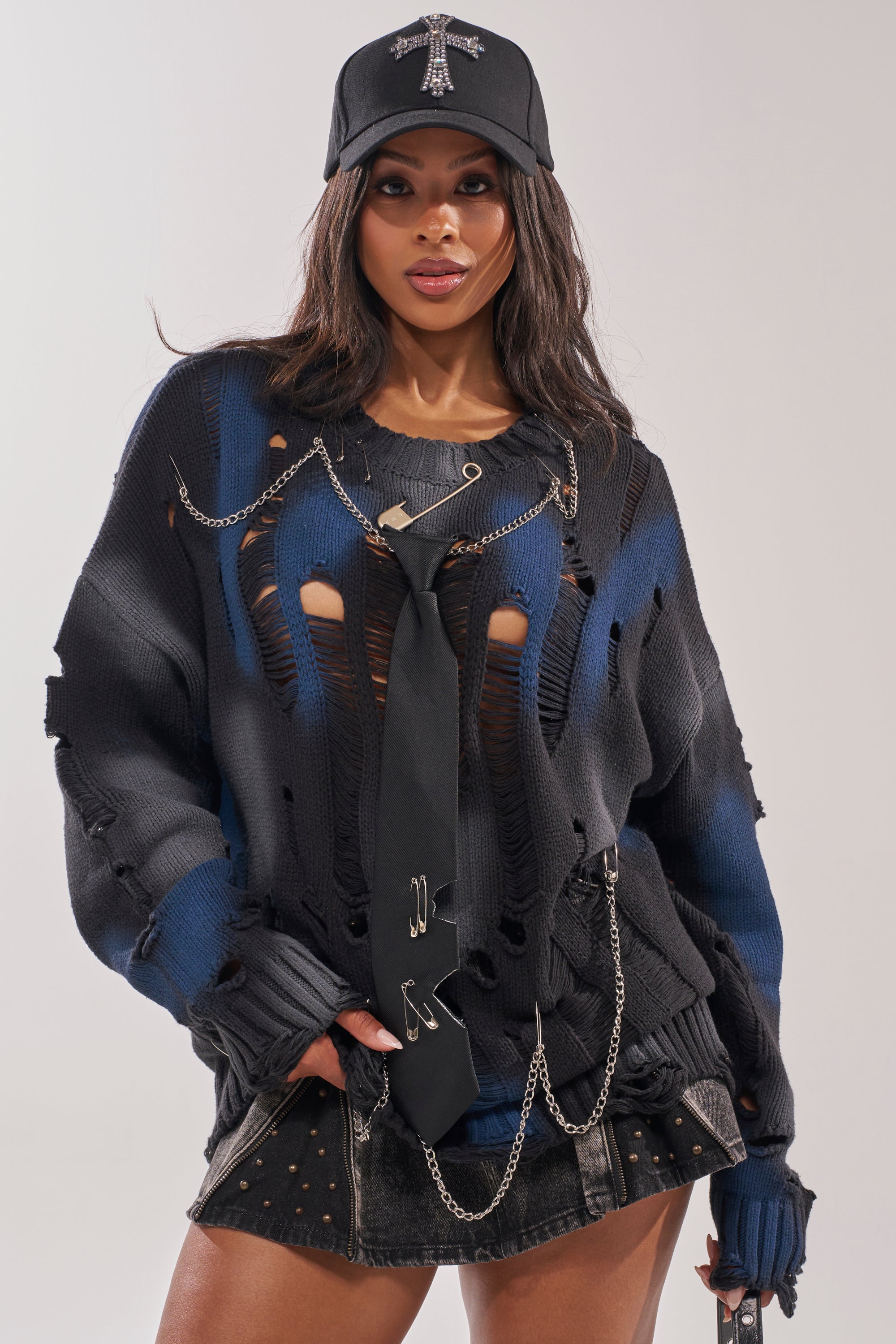 A woman confidently poses against a plain light background, wearing the RAWR DISTRESSED OVERSIZED SWEATER styled with a black cap, silver chains, a black tie, and a short black skirt.