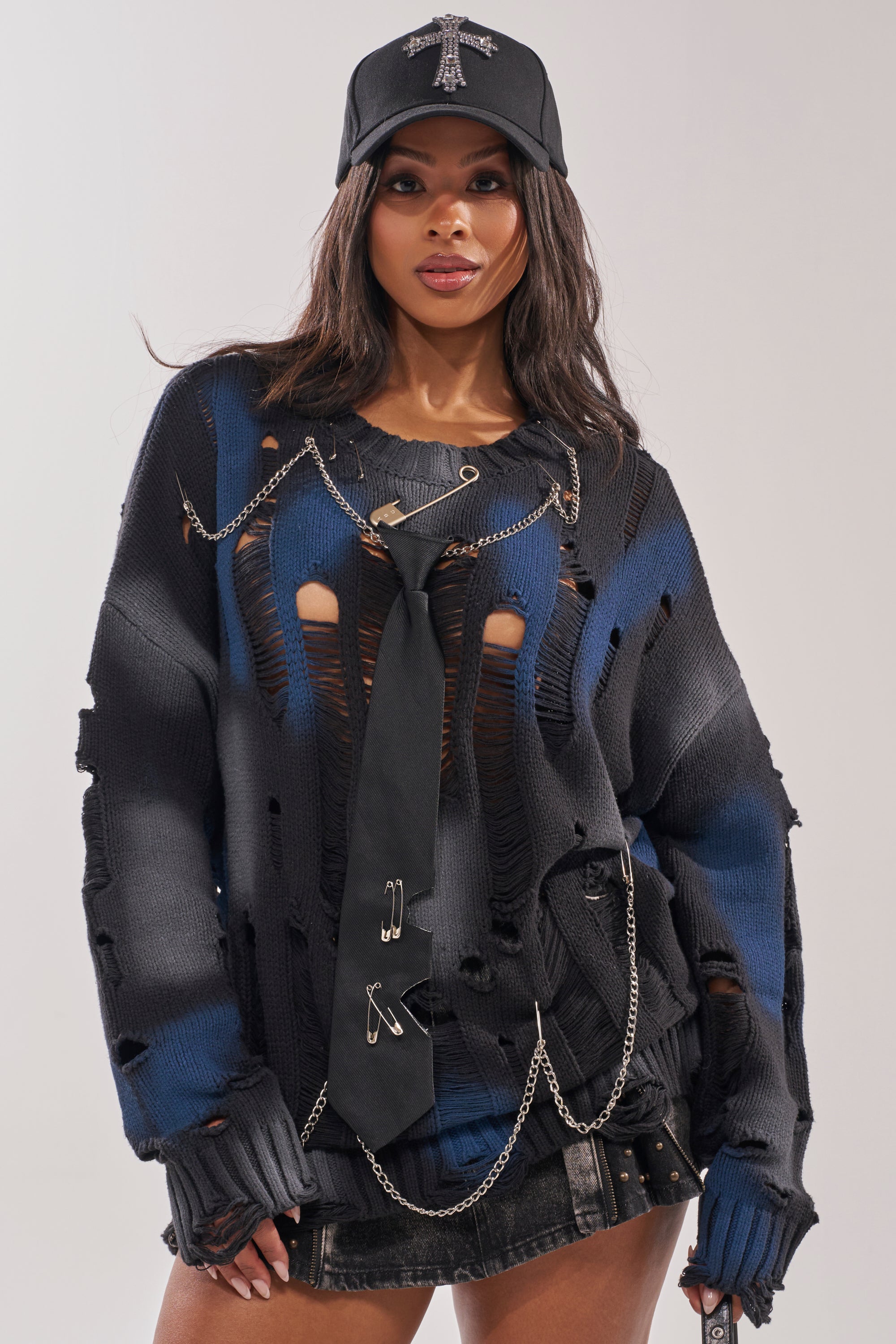 A woman wears the RAWR DISTRESSED OVERSIZED SWEATER, paired with a black tie with safety pins, a black denim mini skirt, a rhinestone cross black cap, and decorative chains for a bold street-style look.