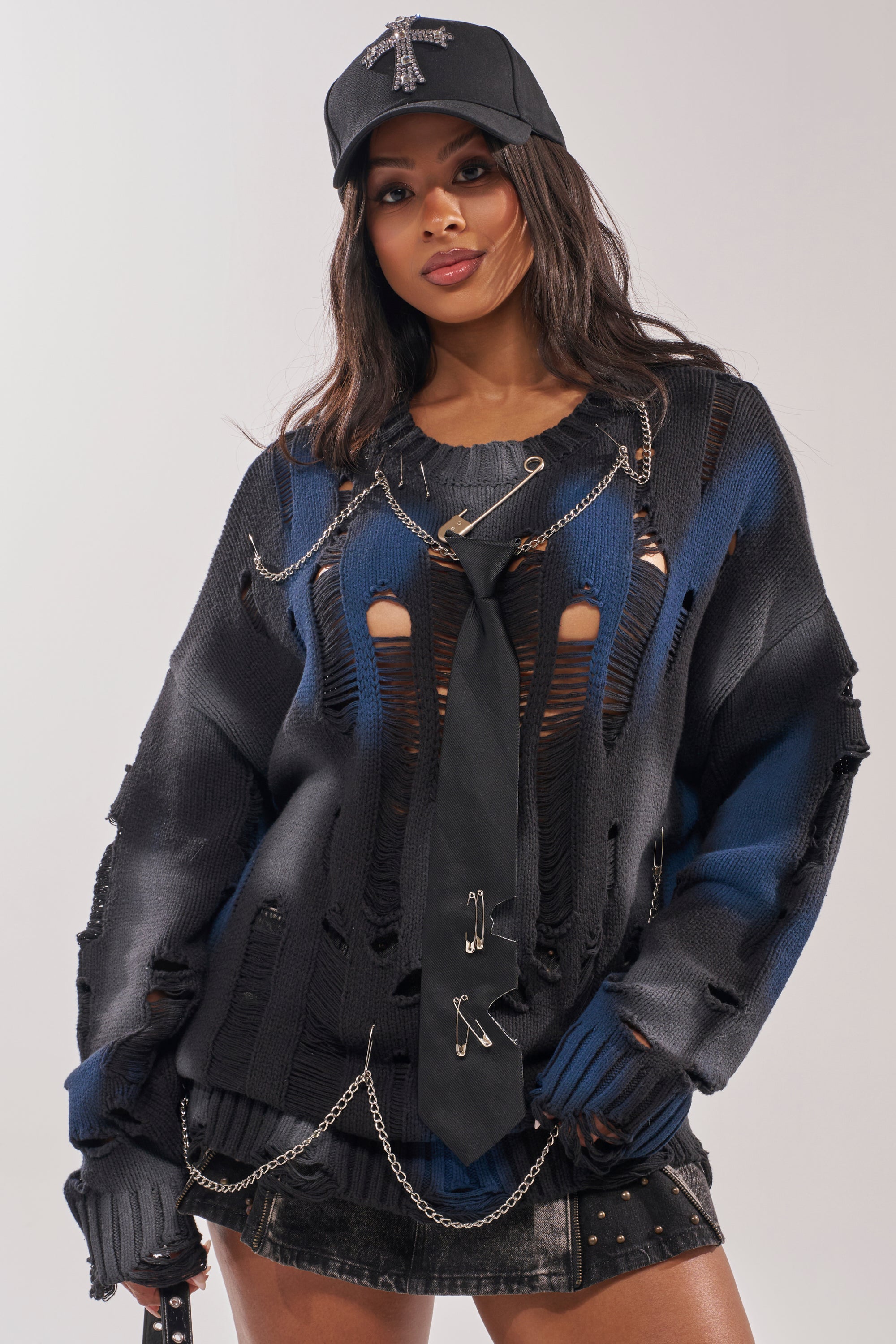 A woman poses confidently against a plain background in a black cap, black tie, studded short skirt, and the RAWR DISTRESSED OVERSIZED SWEATER—punk-inspired with stripes, holes, and safety pins.