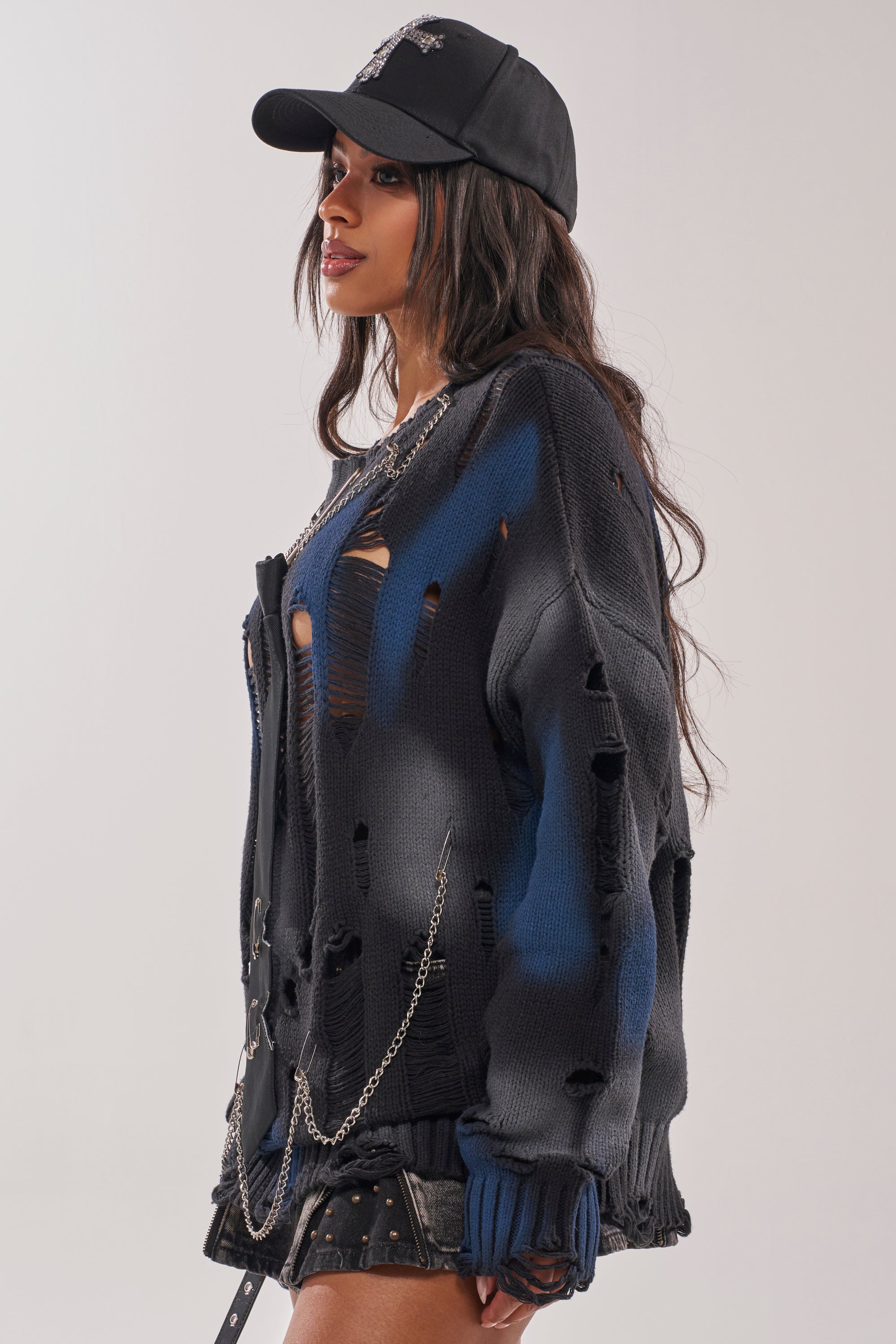 Woman wearing the RAWR DISTRESSED OVERSIZED SWEATER with holes, styled over a blue top, black mini skirt with studs, chain accessories, and a black cap, posing against a plain background.