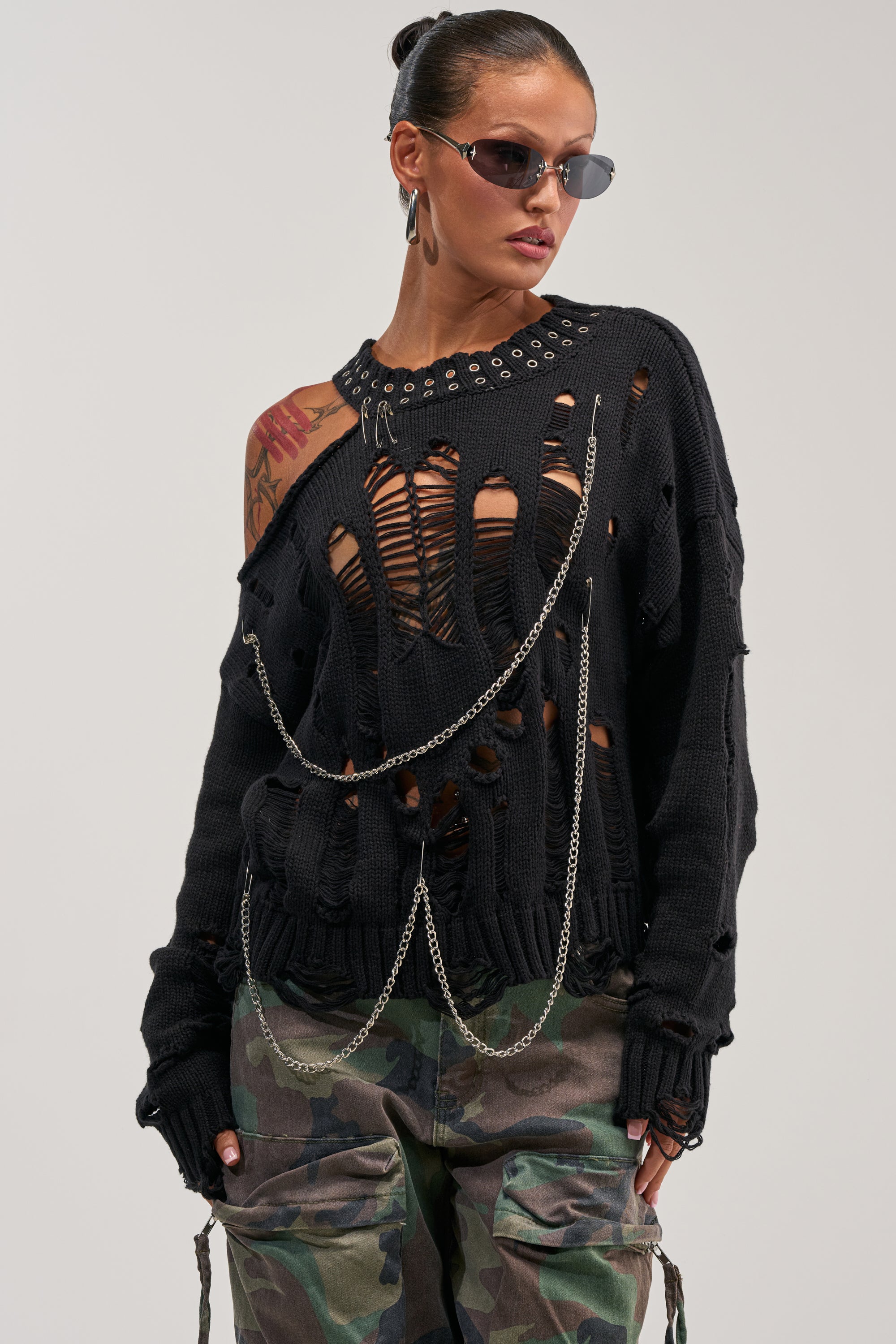 A woman models the CHOPPED UP DISTRESSED SWEATER, pairing it with camo pants, sunglasses, and bold makeup. Her arm tattoos and tied-back hair complete a striking streetwear look.
