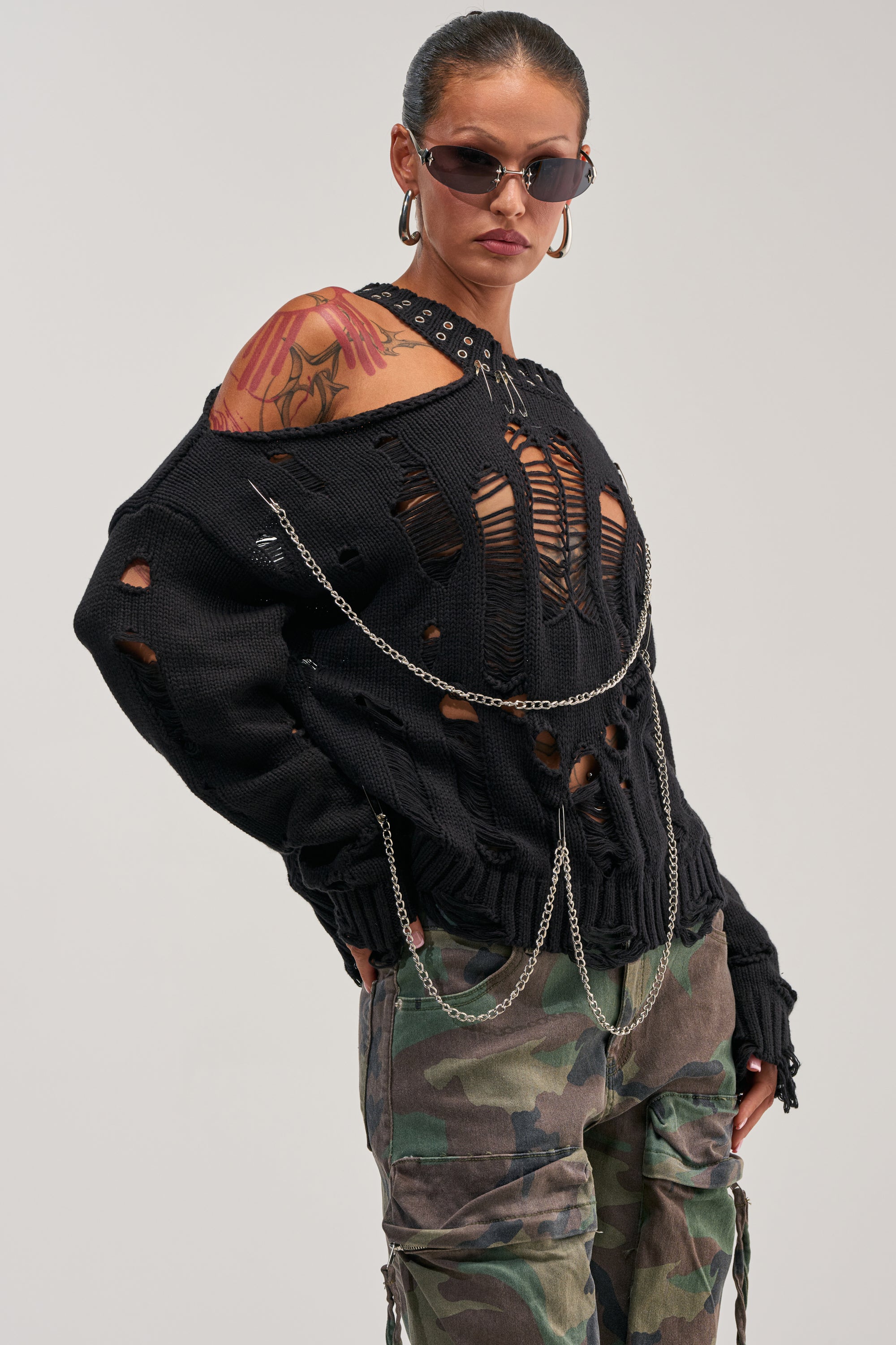 A woman wearing sunglasses and the CHOPPED UP DISTRESSED SWEATER—featuring black distressed details and metal chains—pairs it with camouflage pants and shows off a visible arm tattoo, posing confidently against a plain background.