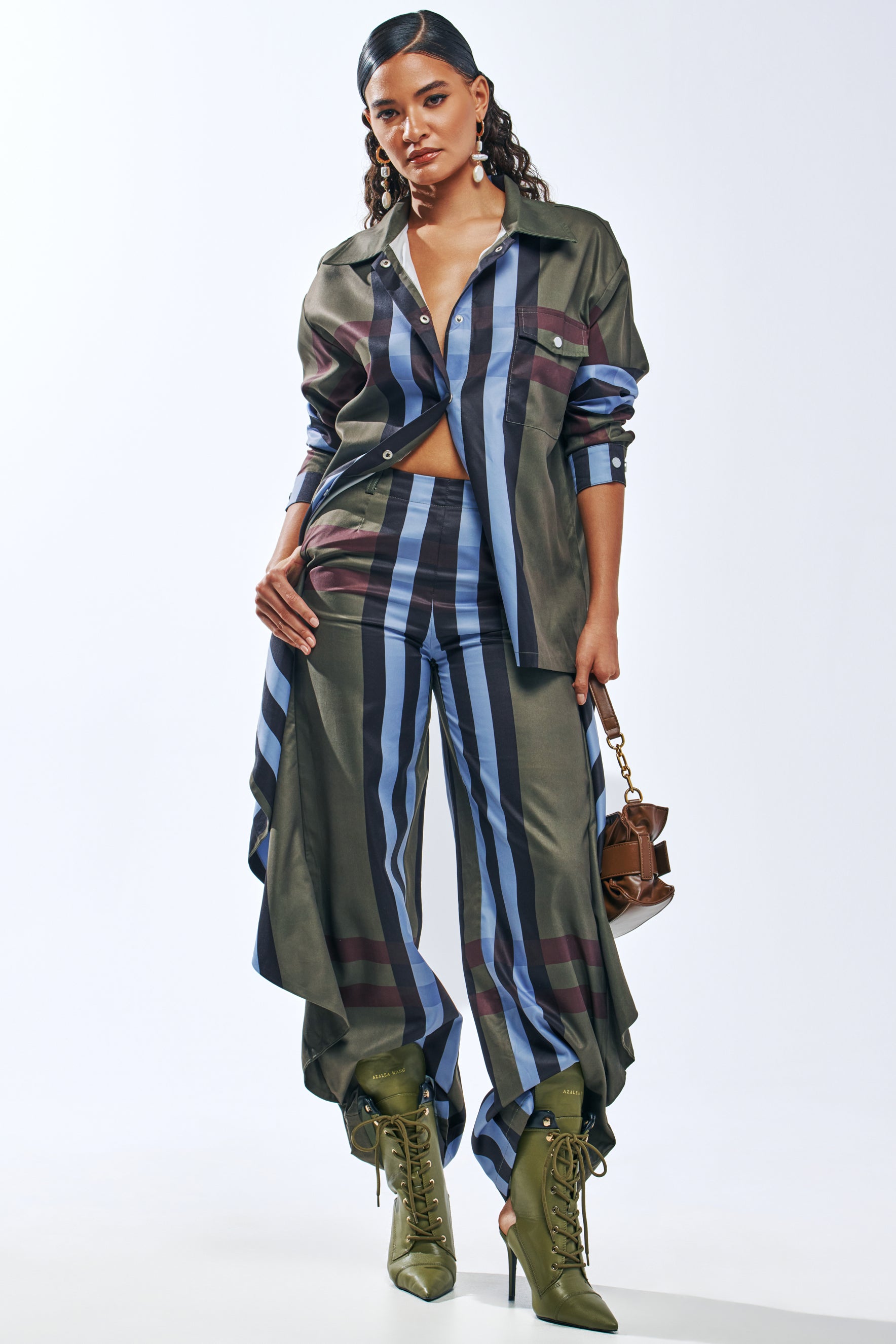 A woman wears a green, blue, and burgundy LUXURY LIFE PLAID TROUSER IN GREEN with a matching shirt, styled with olive green lace-up heeled boots and a small brown handbag.