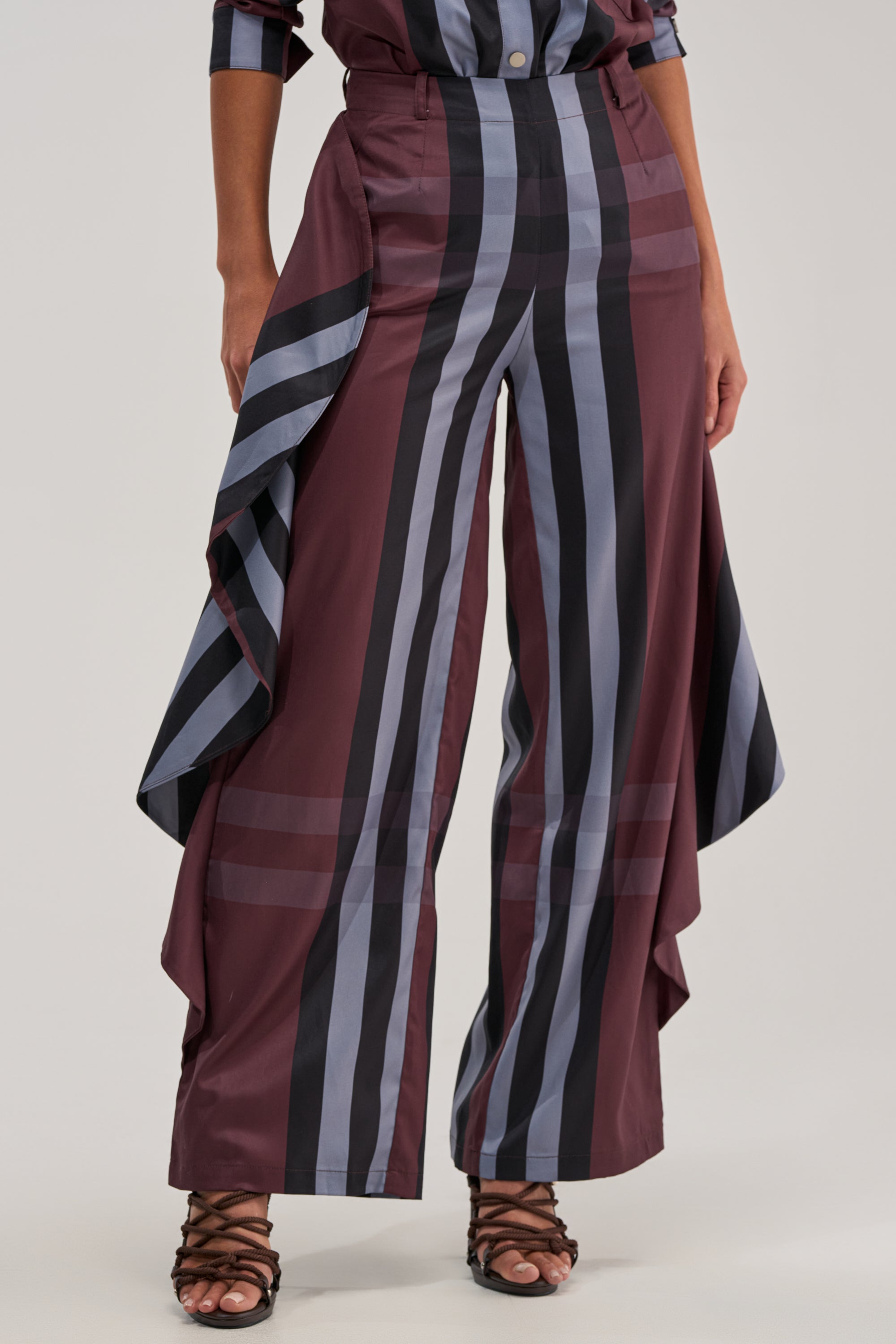 A person wears the LUXURY LIFE PLAID PALAZZO PANT featuring a high-rise fit, wide legs, bold black, gray, and burgundy plaid, and dramatic side drapes, styled with open-toed black sandals.