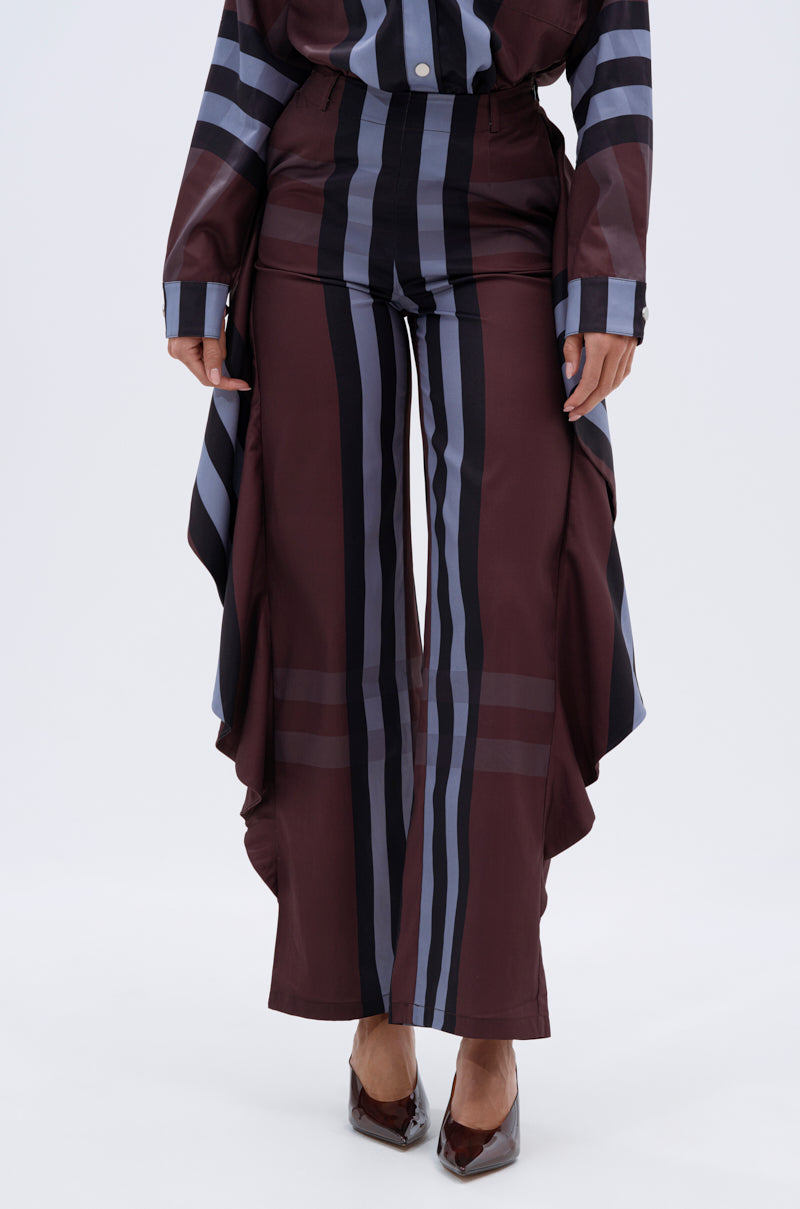 A person wears LUXURY LIFE PLAID PALAZZO PANT featuring vertical black and gray stripes with ruffled sides, paired with a matching long-sleeve top and pointed-toe heels. The image is cropped from the shoulders down.