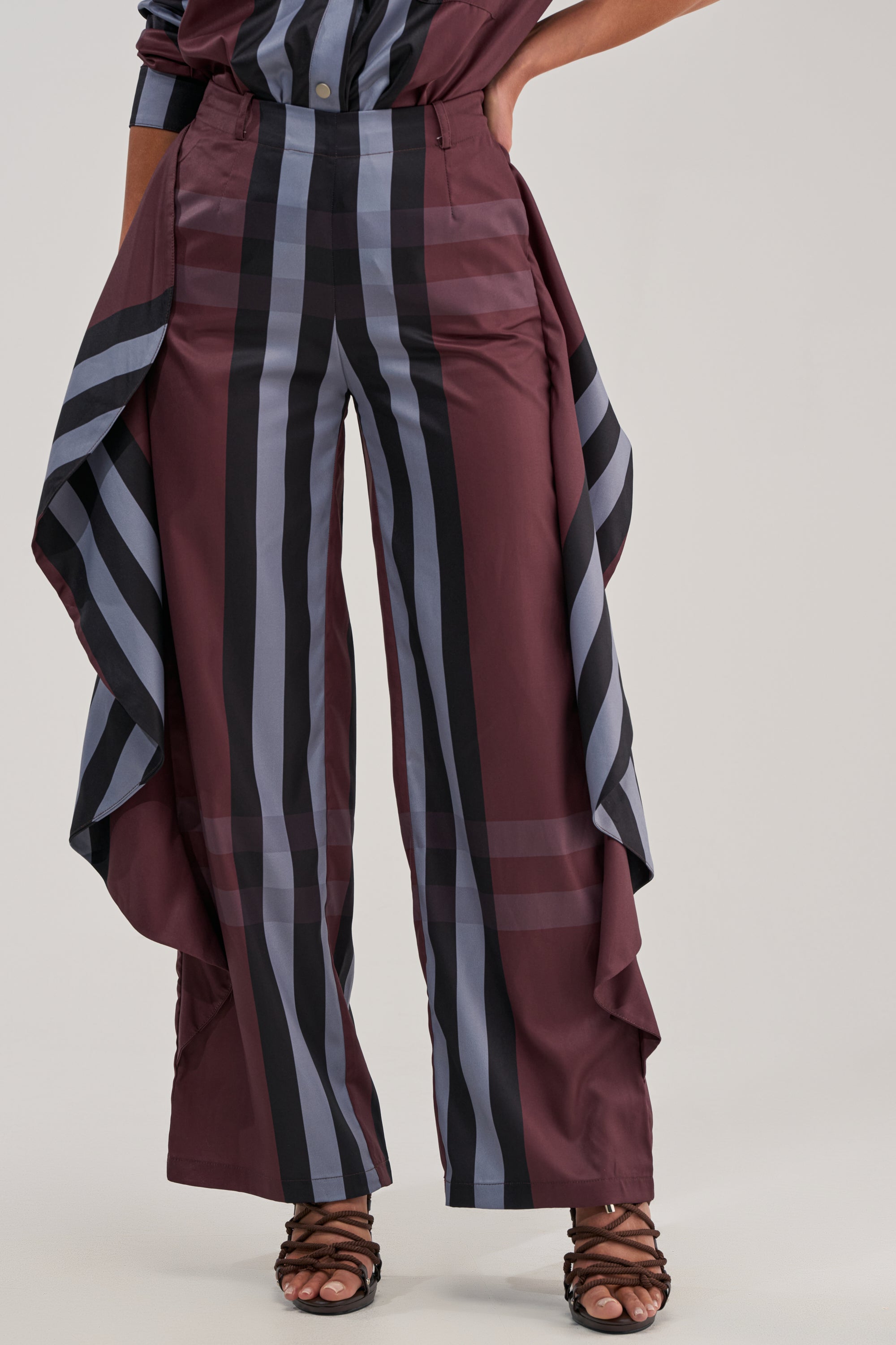 A person wearing the LUXURY LIFE PLAID PALAZZO PANT—high-rise, wide-leg pants with bold burgundy, black, and gray plaid stripes and draped side panels—paired with strappy black heeled sandals.