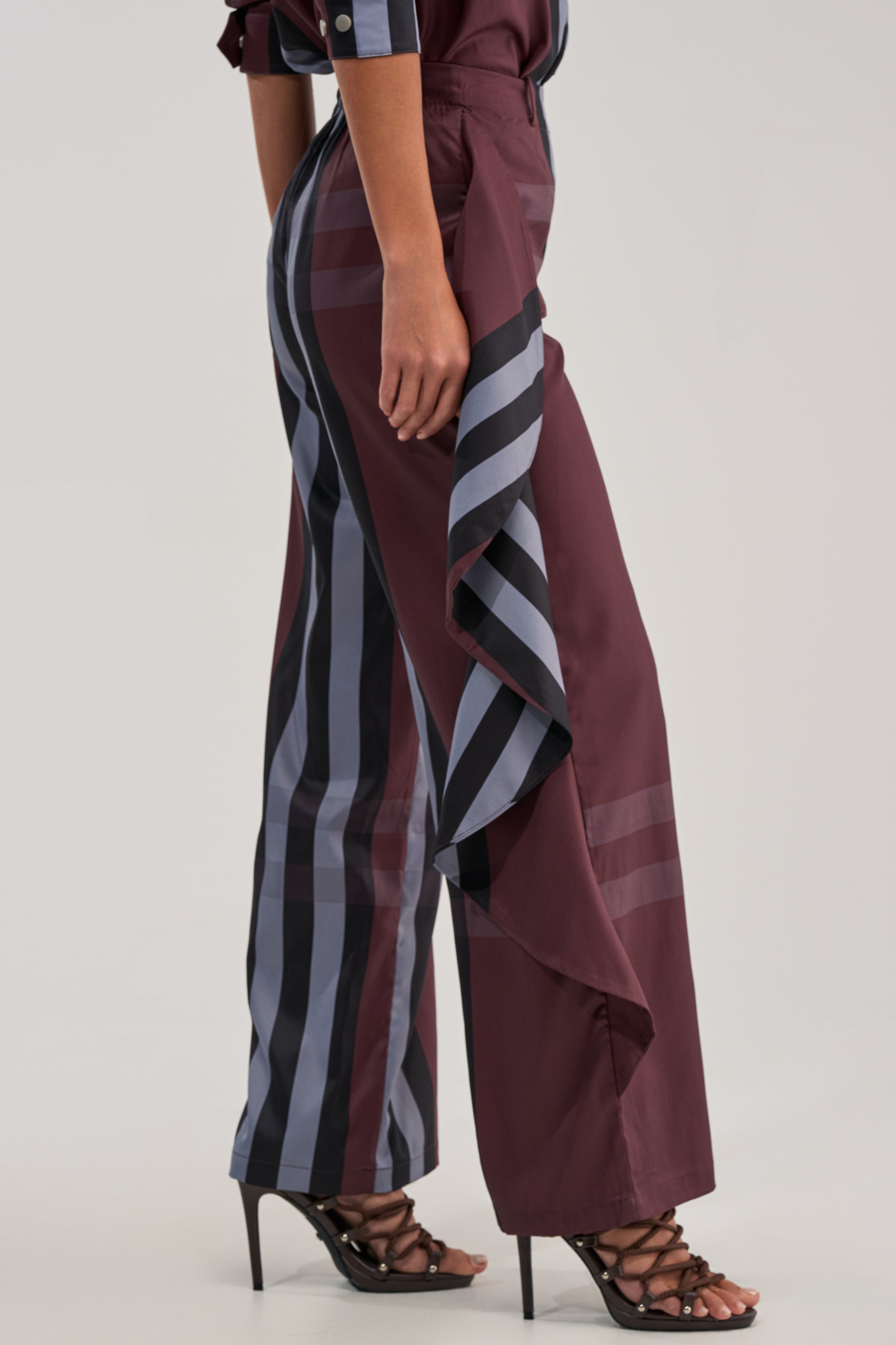 Wearing the LUXURY LIFE PLAID PALAZZO PANT with navy and burgundy stripes, wide-leg fit, and ruffled sides, paired with strappy high-heeled sandals, a person stands against a light gray background.
