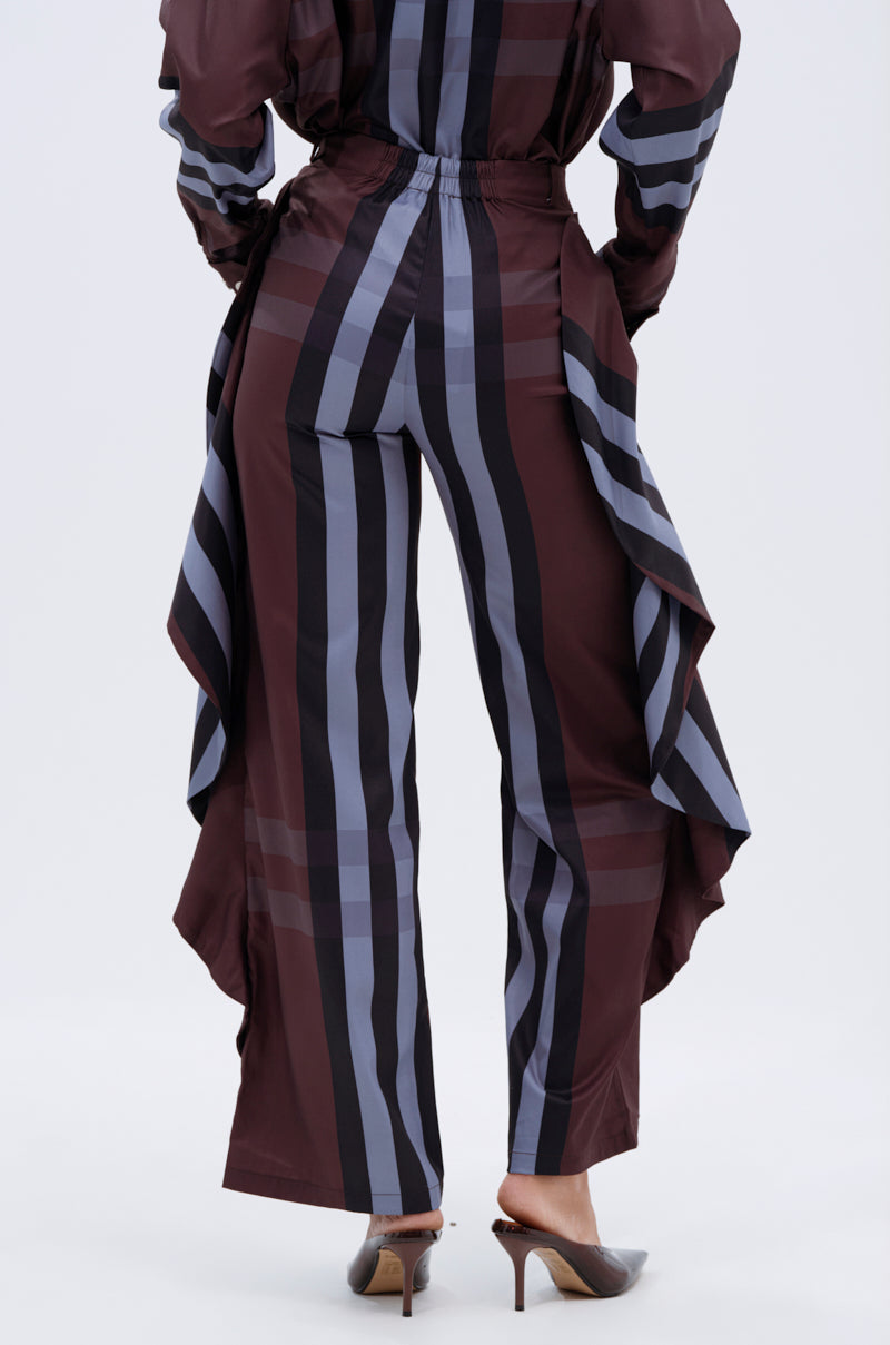 A person stands facing away, wearing the LUXURY LIFE PLAID PALAZZO PANT with vertical maroon, black, and gray stripes plus dramatic ruffled side panels, paired with heeled pointed-toe shoes.