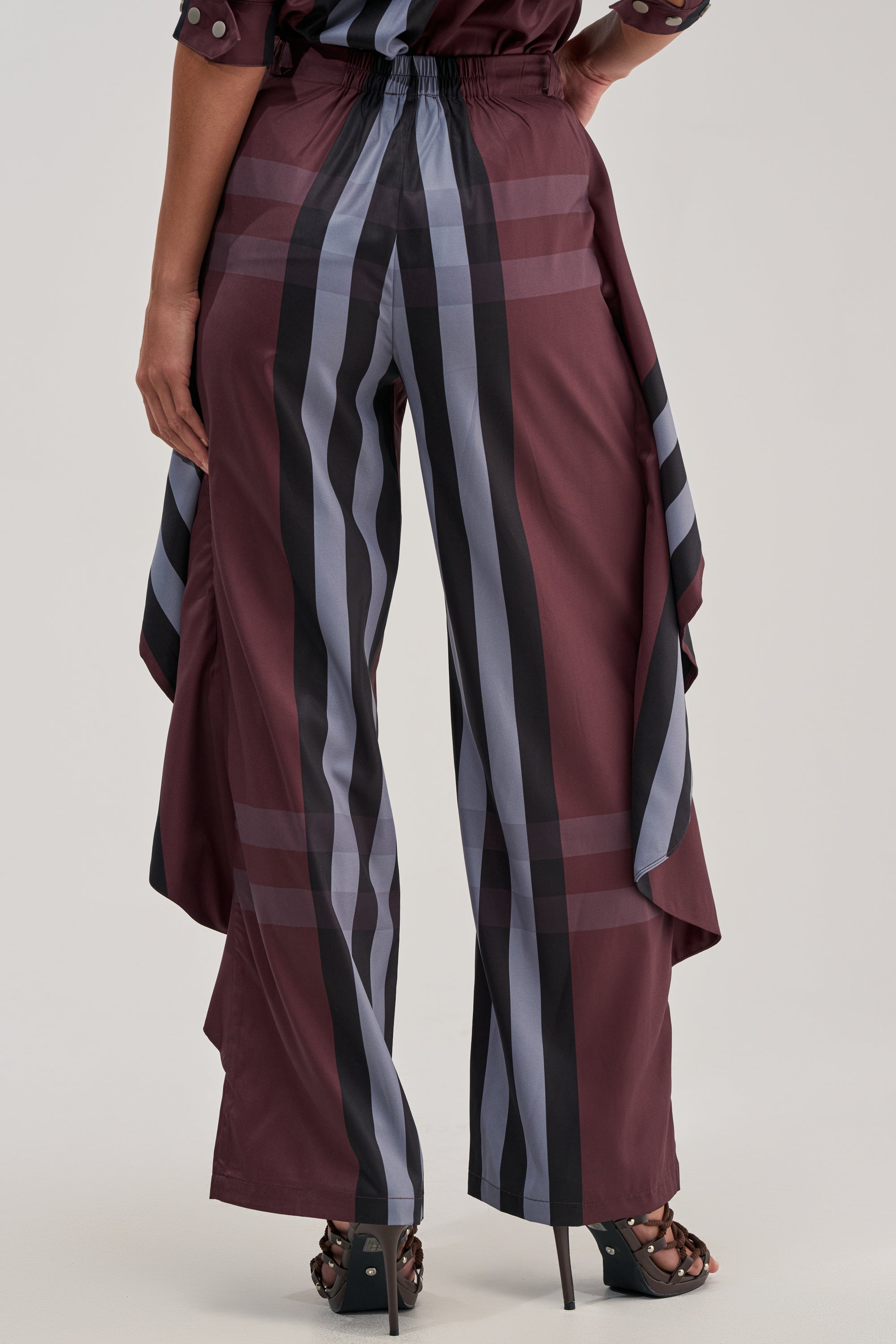 A person stands facing away, wearing the LUXURY LIFE PLAID PALAZZO PANT featuring maroon stripes, a wide-leg silhouette, and draped side panels. They pair it with heeled sandals against a plain, light-colored background.