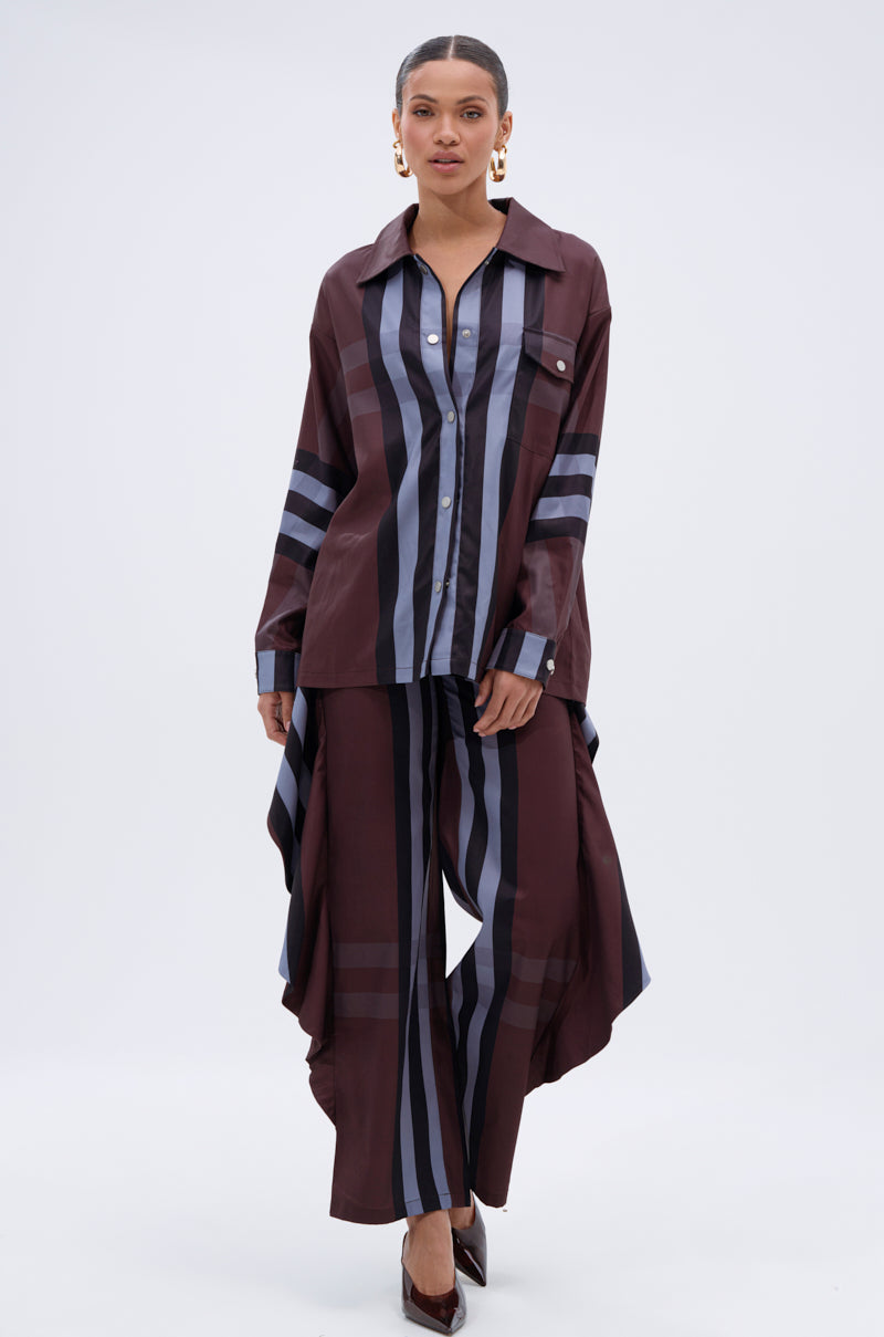 A woman models the LUXURY LIFE PLAID BUTTON DOWN with wide-leg, draped side pants in brown and black stripes, paired with pointed dark heels against a plain light background.