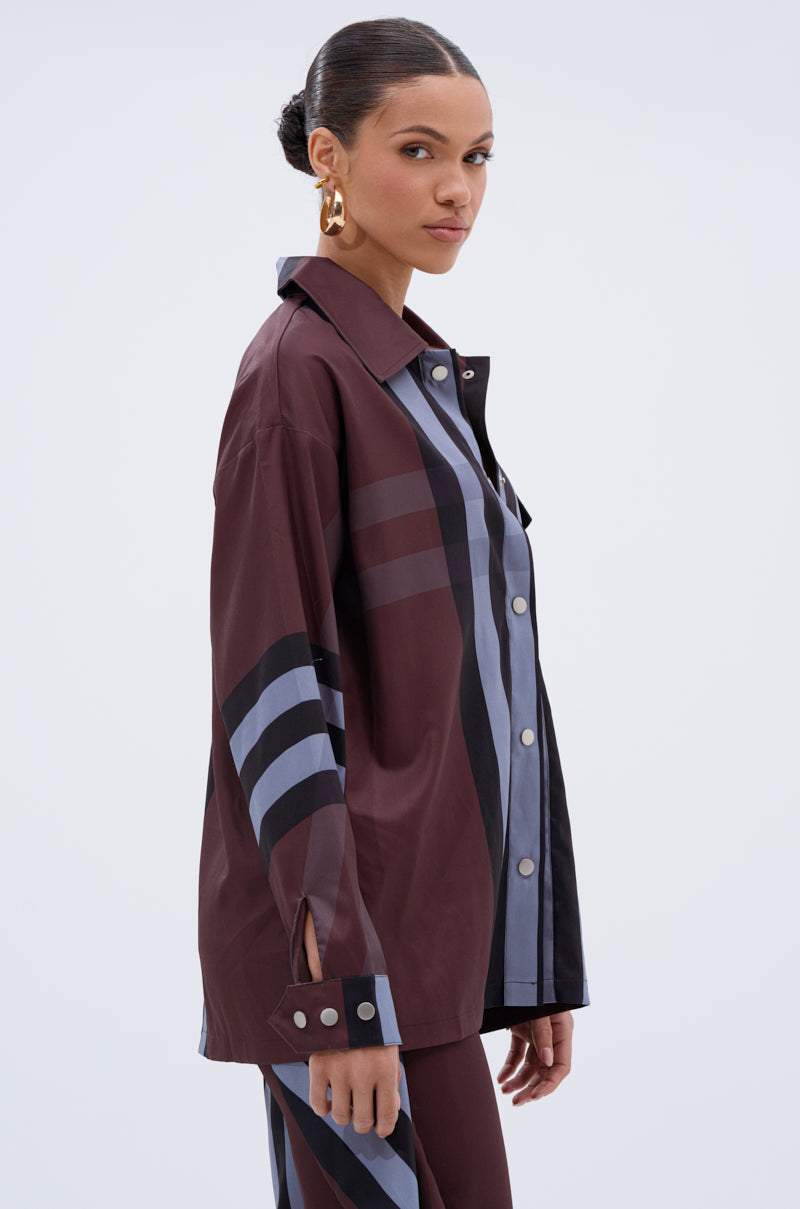 A woman with slicked-back hair and large gold hoop earrings wears the LUXURY LIFE PLAID BUTTON DOWN, a loose shirt with bold burgundy, navy, and black stripes, standing sideways against a plain light background.