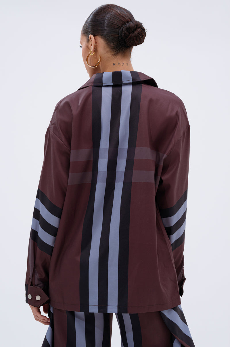 A person with hair in a neat bun, wearing the LUXURY LIFE PLAID BUTTON DOWN in maroon with black and gray stripes, stands facing away. Gold hoop earrings and neck tattoos add bold accents to the look.