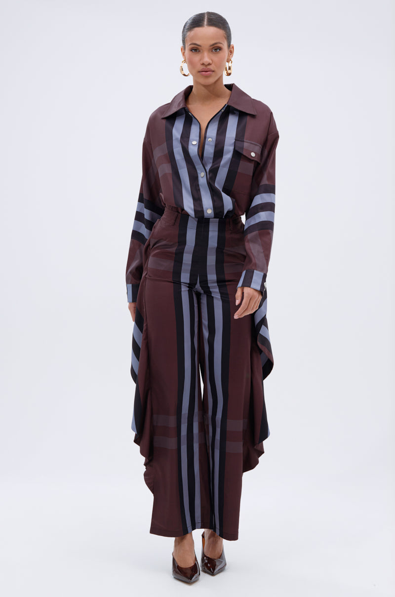 A woman faces forward wearing the LUXURY LIFE PLAID BUTTON DOWN in brown, black, and gray, styled with matching wide-leg pants. The collared long-sleeve shirt features draped sides and is paired with hoop earrings and heels.