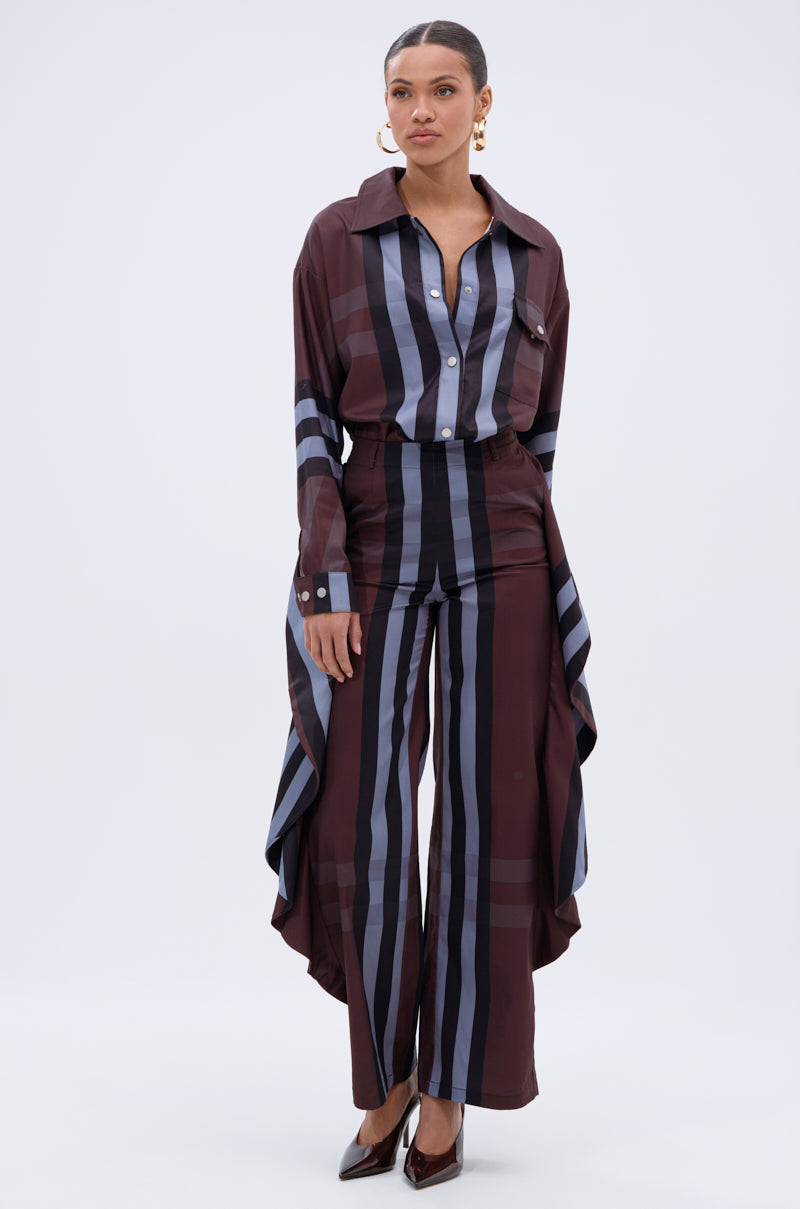 A woman stands against a plain background wearing the AKIRA Label LUXURY LIFE PLAID BUTTON DOWN shirt with matching wide-leg pants, draped fabric panels, pointed heels, and large hoop earrings.