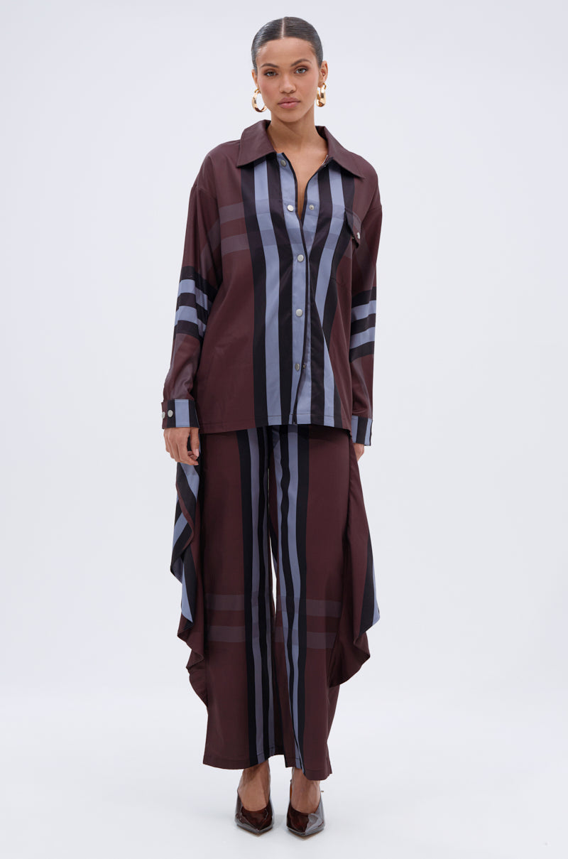 A woman faces forward in the LUXURY LIFE PLAID BUTTON DOWN, a loose burgundy shirt paired with matching wide-leg pants featuring black and gray vertical stripes. She accessorizes with gold hoop earrings, slicked-back hair, and pointed dark heels.