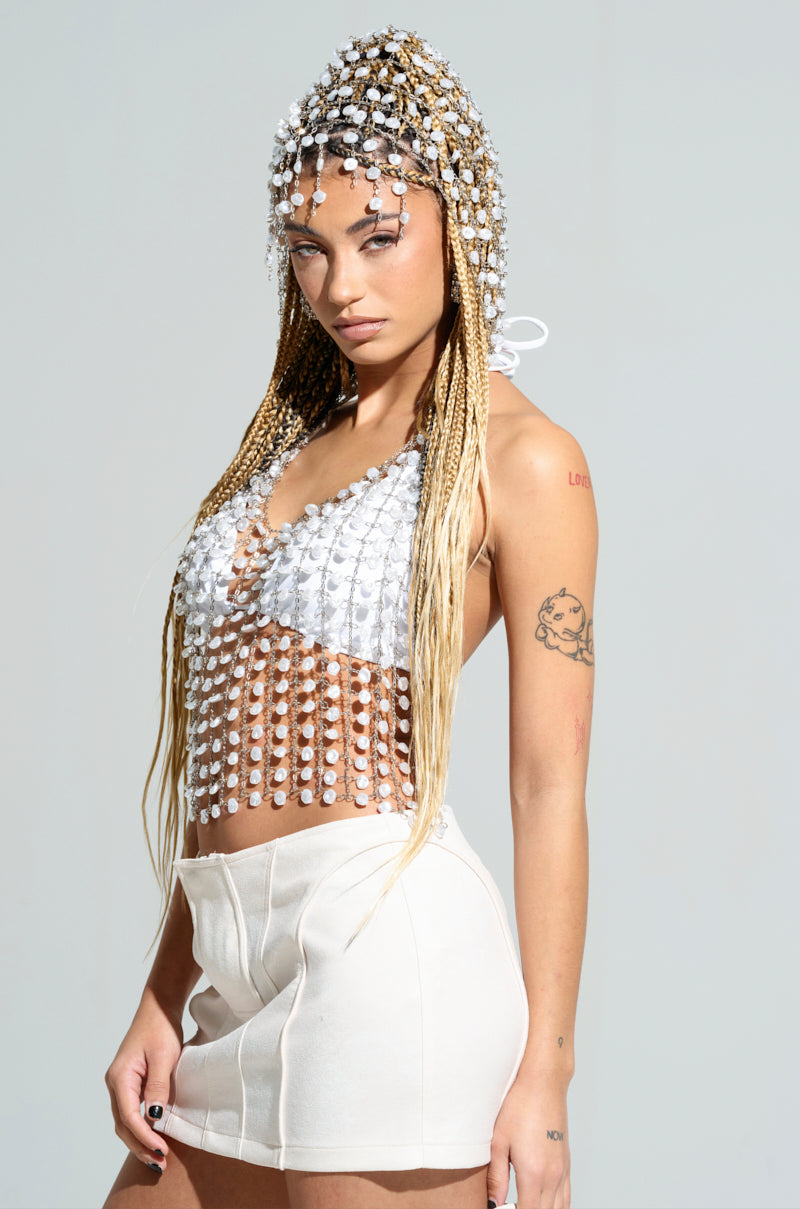A person with long braids and a pearl-adorned headpiece stands confidently, wearing the "Just Like Heaven Hooded Chain Top," metallic chain accessories, and a white skirt. They display tattoos on their arms against a plain, neutral background.