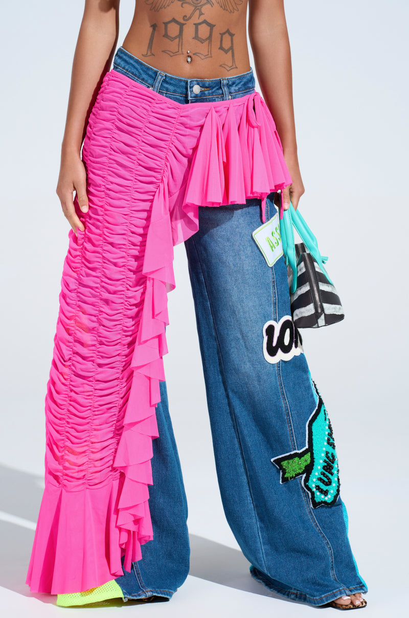 A person wears wide-leg denim jeans with colorful patches and the LAYERED UP MESH RUFFLE BELT SKIRT in bright pink draped over one leg, accessorized with a small green purse, striped bag, and a "1979" torso tattoo.