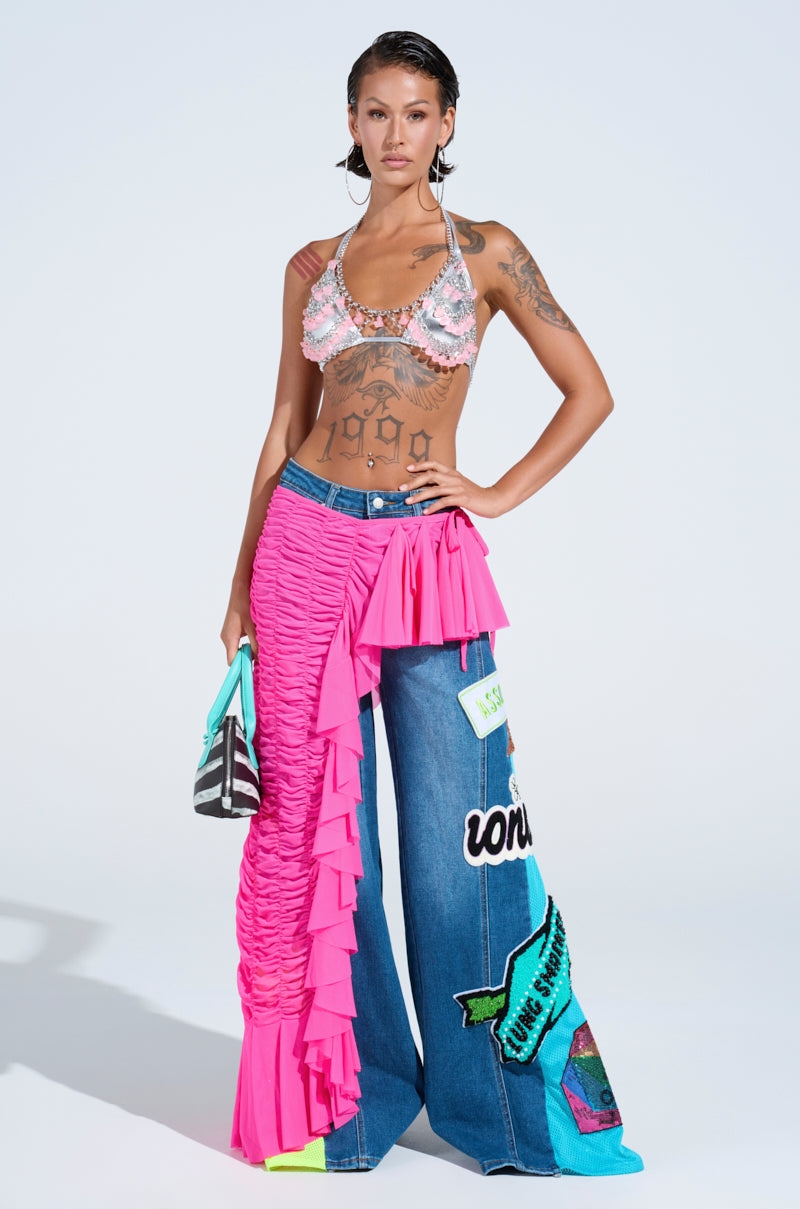A person stands confidently in festival fashion, wearing a beaded bikini top and the LAYERED UP MESH RUFFLE BELT SKIRT over one leg, paired with wide-leg blue jeans featuring colorful patches and text. They carry a striped bag and show off arm and torso tattoos.