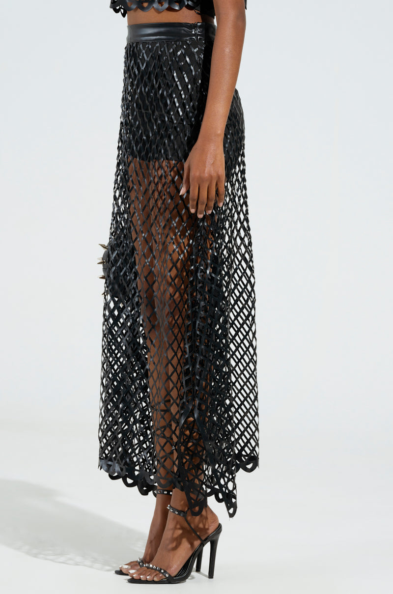A person dressed in a fashionable ensemble featuring the BREAKING THROUGH MAXI SKIRT with its cage-inspired mesh design and high-heeled shoes is standing against a plain background. The skirt showcases a layered look, while the heels boast clear material with black detailing.