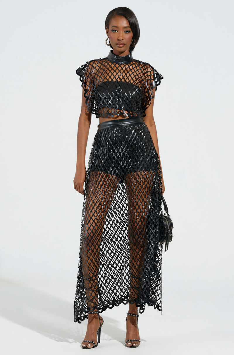 A woman showcases a stylish black netted ensemble, wearing a cage-inspired crop top paired with the BREAKING THROUGH MAXI SKIRT that features a faux leather structure and shorts underneath. She completes her look with high heels and a small handbag, set against a simple light background.