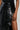 Close-up of a person wearing the KEEP WALKING MIDI SKIRT, a black high-rise design featuring an asymmetrical front panel. Their hand rests on their hip, highlighting the glossy texture of the faux leather material.