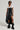 A stylish person wearing sunglasses, a white and black top, a black corset, the KEEP WALKING MIDI SKIRT with its asymmetrical high-rise faux leather design, and unique black boots poses confidently against a plain background.