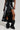 A person models the KEEP WALKING MIDI SKIRT in black faux leather, paired with black slouchy boots adorned with metal studs. The outfit creates a dramatic contrast between the skirt's elegant design and the boots' rugged look.