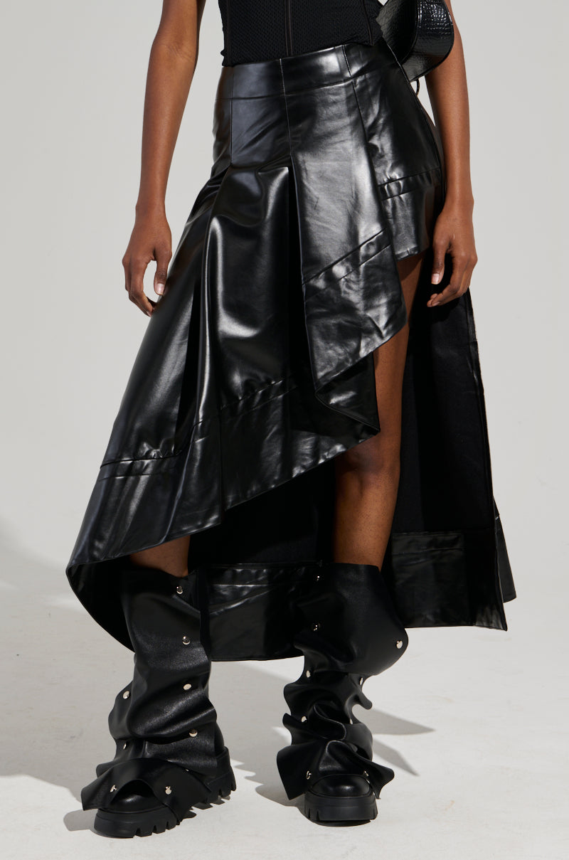 A person wearing the KEEP WALKING MIDI SKIRT, a black asymmetrical faux leather skirt, paired with high-rise black boots stands against a light gray background, showcasing an edgy and bold fashion style.