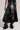 A person wearing the KEEP WALKING MIDI SKIRT, a black asymmetrical faux leather skirt, paired with high-rise black boots stands against a light gray background, showcasing an edgy and bold fashion style.