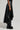 A person wearing the KEEP WALKING MIDI SKIRT, a black faux leather piece with a high-rise fit and asymmetrical hem, stands against a plain background with black studded boots. Their hand is relaxed by their side.